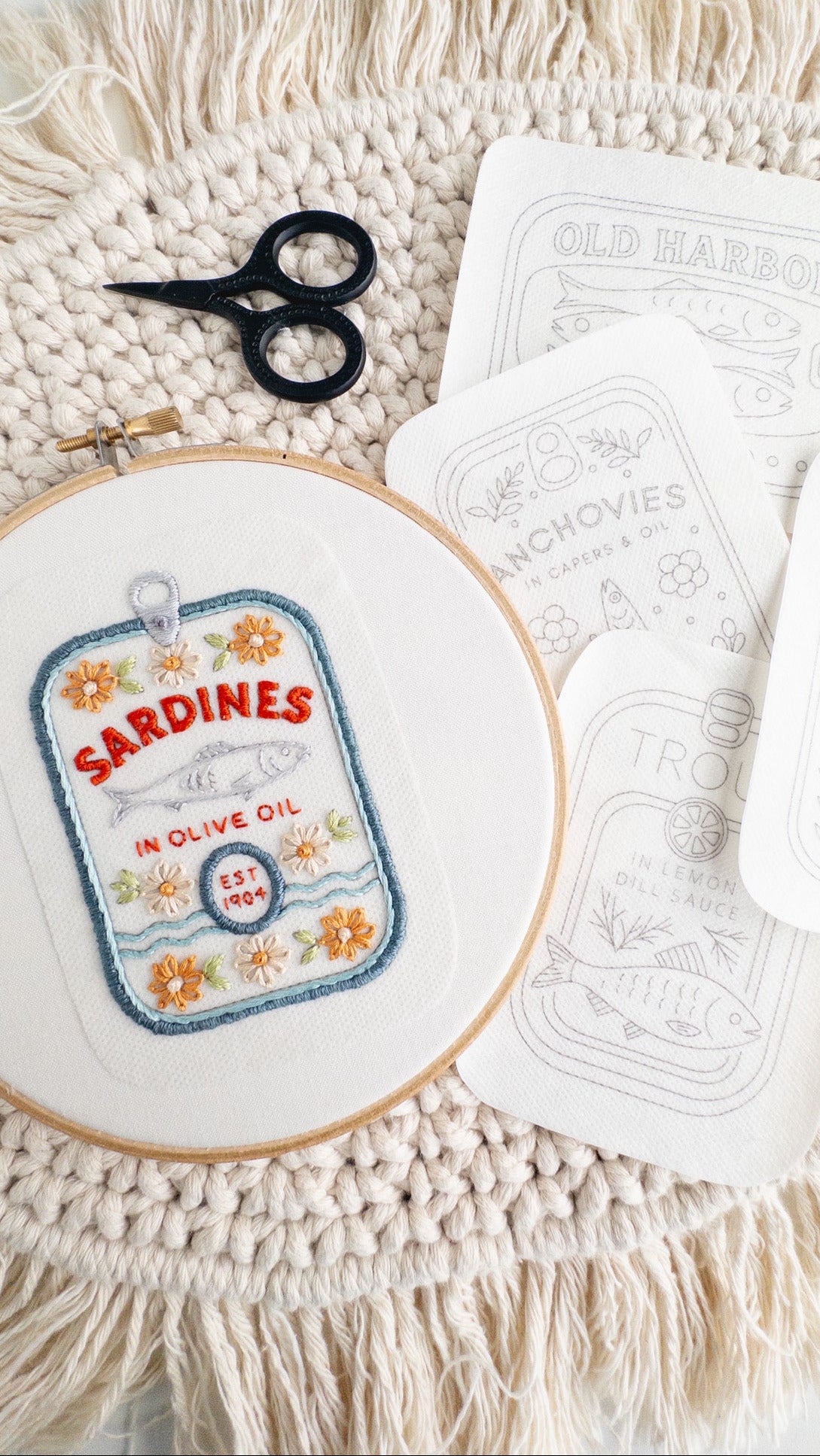 Embroidery hoop with 'Sardines in Olive Oil' design, scissors, and pattern paper on a textured surface.