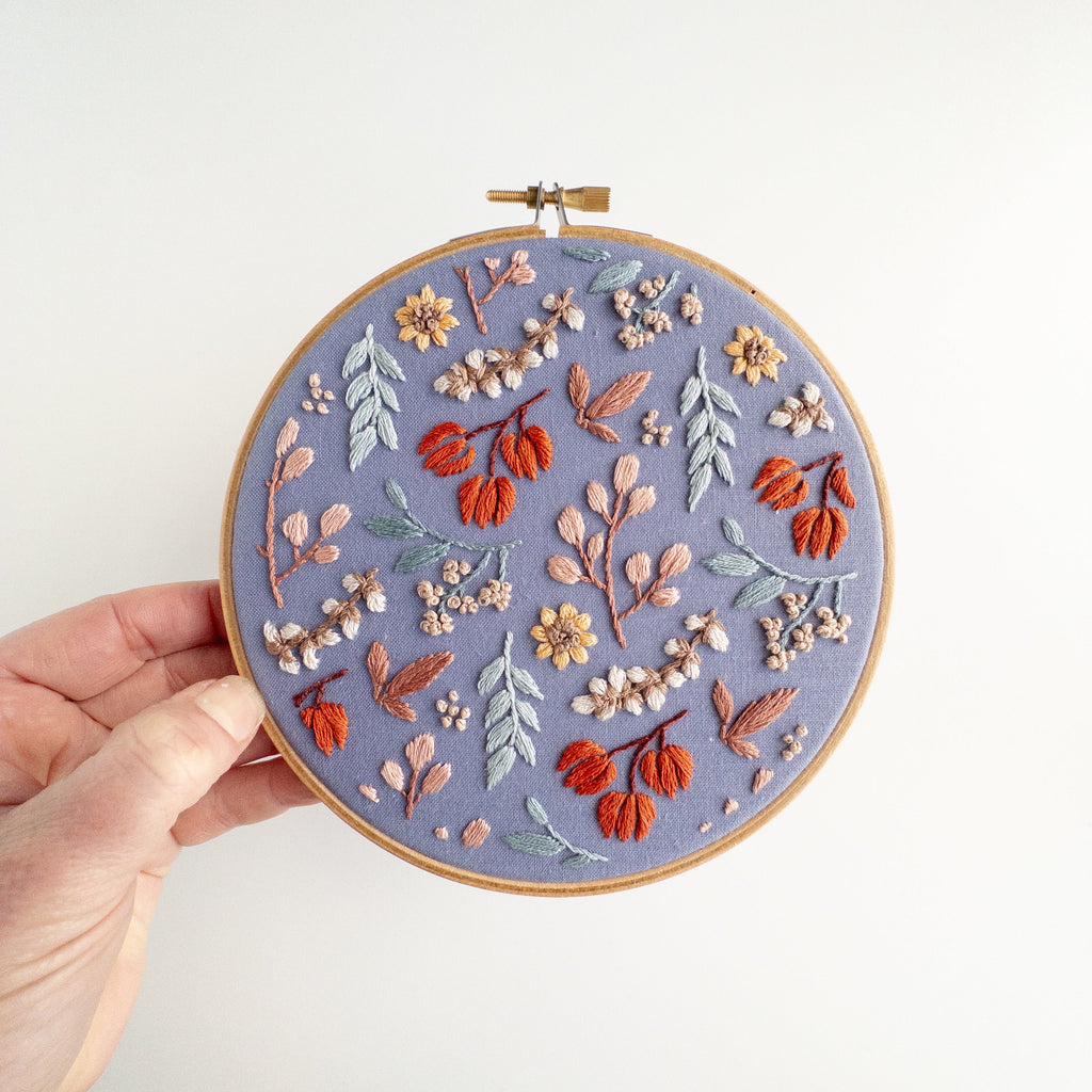 Hand holding a 6-inch embroidery hoop with a "Twilight Meadow" fall floral pattern on moody fabric.
