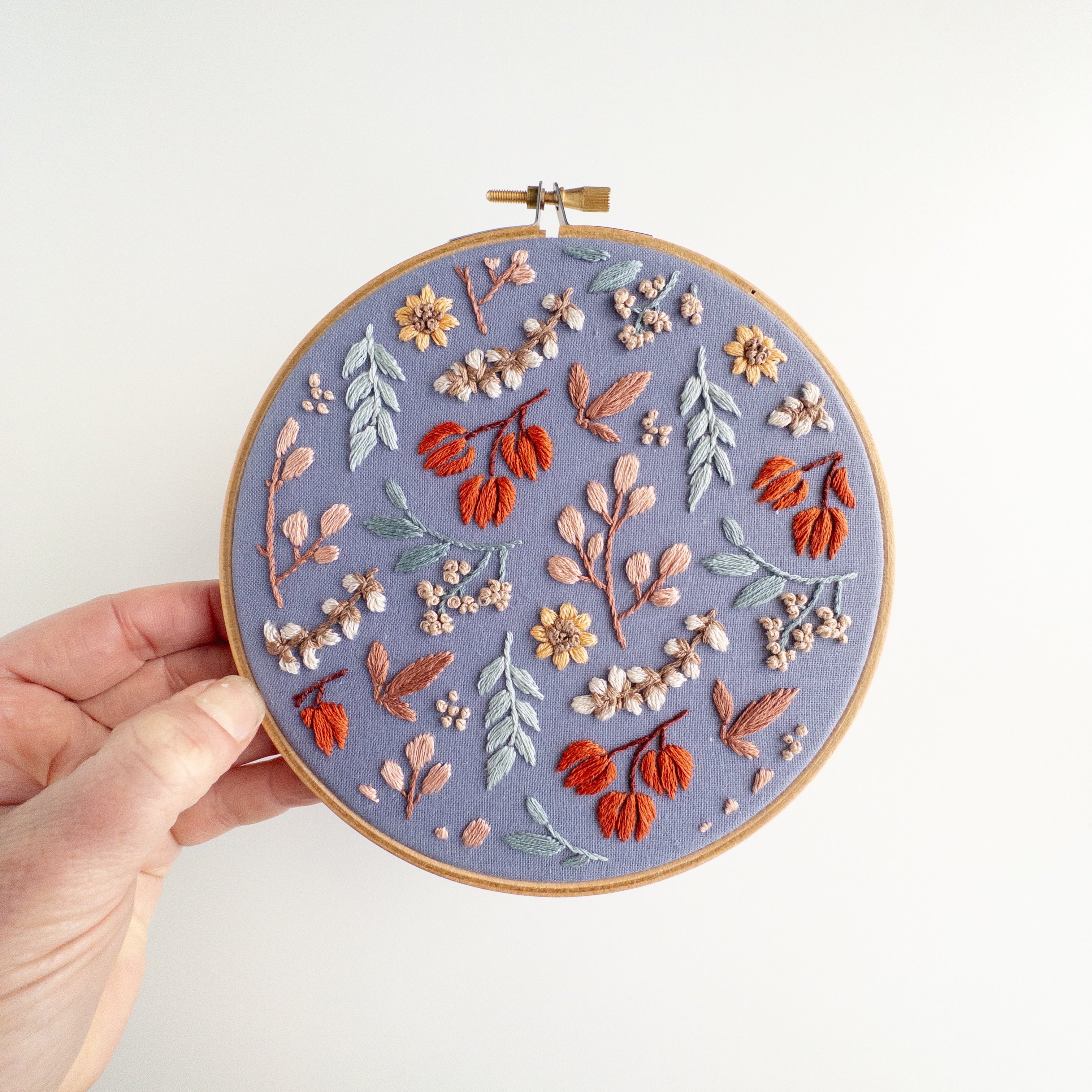 Hand holding a 6-inch embroidery hoop with a "Twilight Meadow" fall floral pattern on moody fabric.