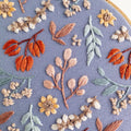 Twilight Meadow embroidery: fall floral design with sunflowers, wild cotton, and leaves on moody blue fabric in a 6-inch hoop.
