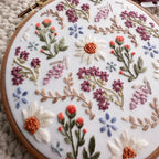 Embroidery hoop with a wildflower field design featuring daisies, lavender, and various floral stitches on white fabric.