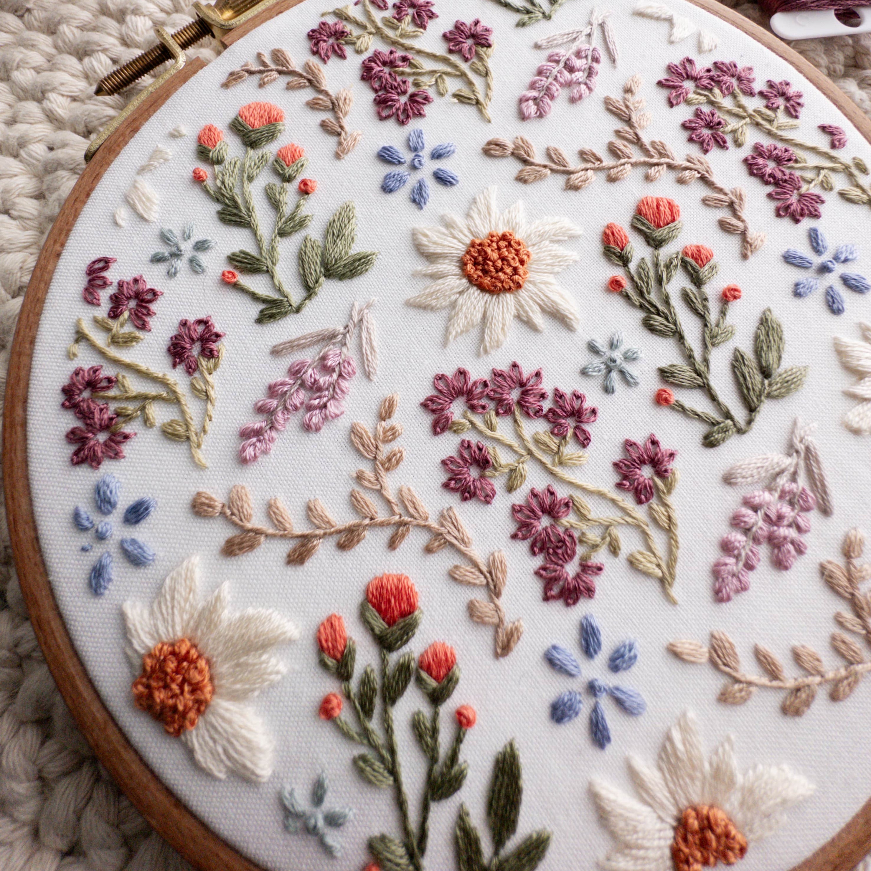 Embroidery hoop with a wildflower field design featuring daisies, lavender, and various floral stitches on white fabric.