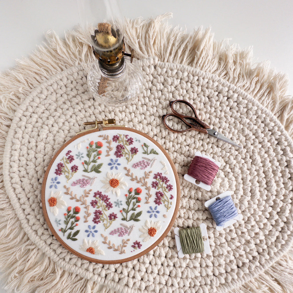 Embroidered wildflower field design in a 6-inch hoop with daisies, lavender, and colorful threads on a macrame mat.