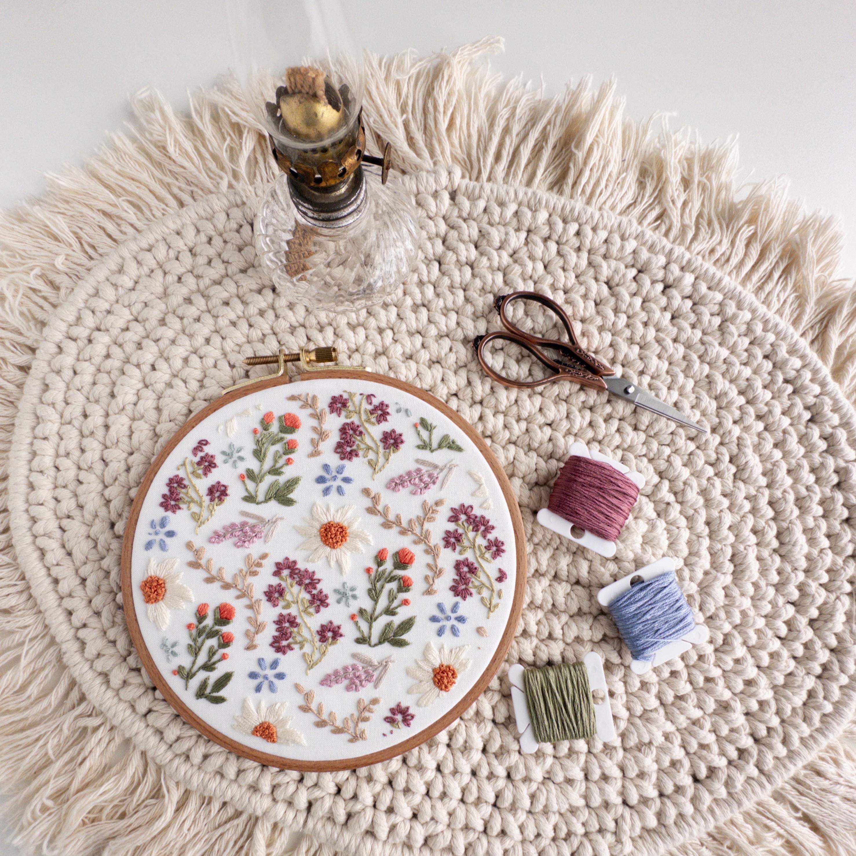 Embroidered wildflower field design in a 6-inch hoop with daisies, lavender, and colorful threads on a macrame mat.
