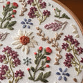 Close-up of a wildflower embroidery pattern with colorful daisies, lavender, and floral designs on white fabric in a hoop.