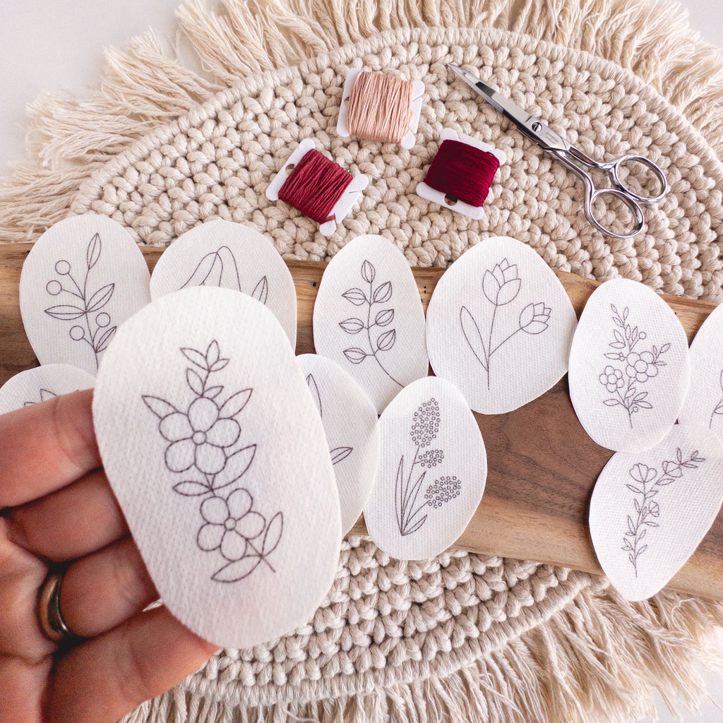 Hand holding a wildflower stick and stitch embroidery pattern, with other patterns, floss, and scissors on a macrame mat.